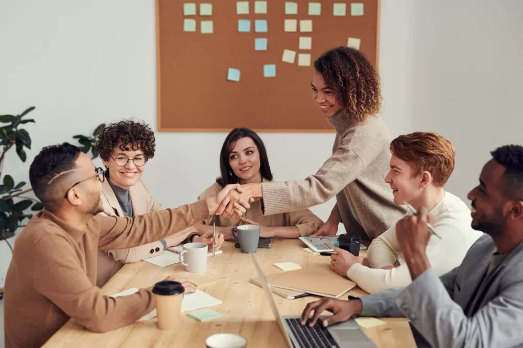 Potential of your people. Group of diverse individuals sitting around a boardroom table with coffee mugs, notebooks and laptop. A Black woman is reaching out across the table with her hand extended shaking the hand of a Black man.